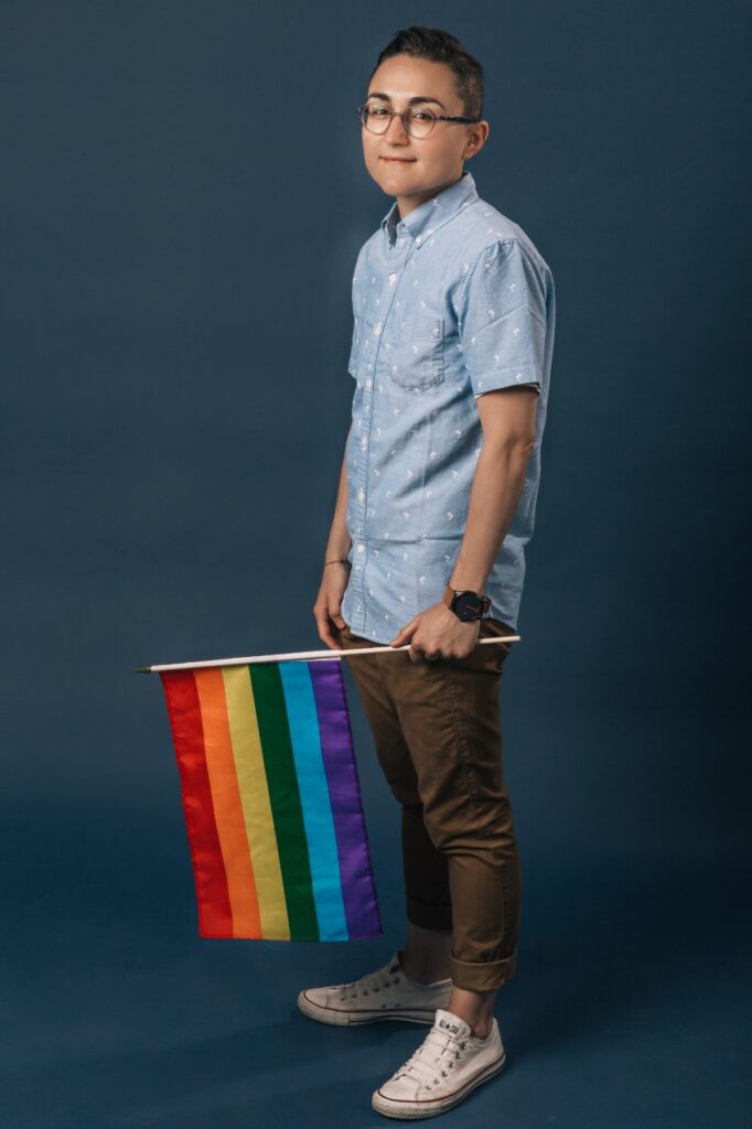 Persona dai capelli corti, gli occhiali da vista, camicia azzurra fuori dai pantaloni kaki. In mano ha una bandiera rainbow.